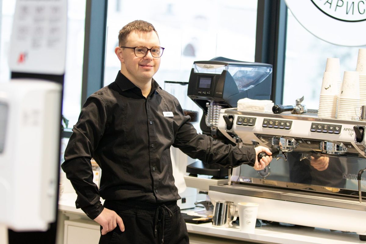 A barista in a black shirt smiles beside an espresso machine.