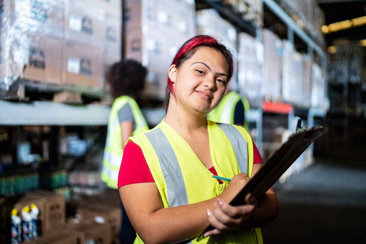 A young woman in a safety vest holds a clipboard in a warehouse.