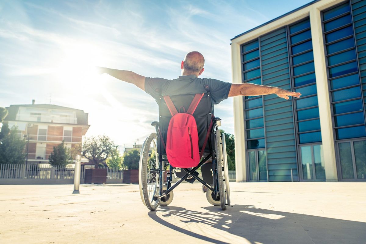 A man in a wheelchair spreads his arms wide outside an office building.