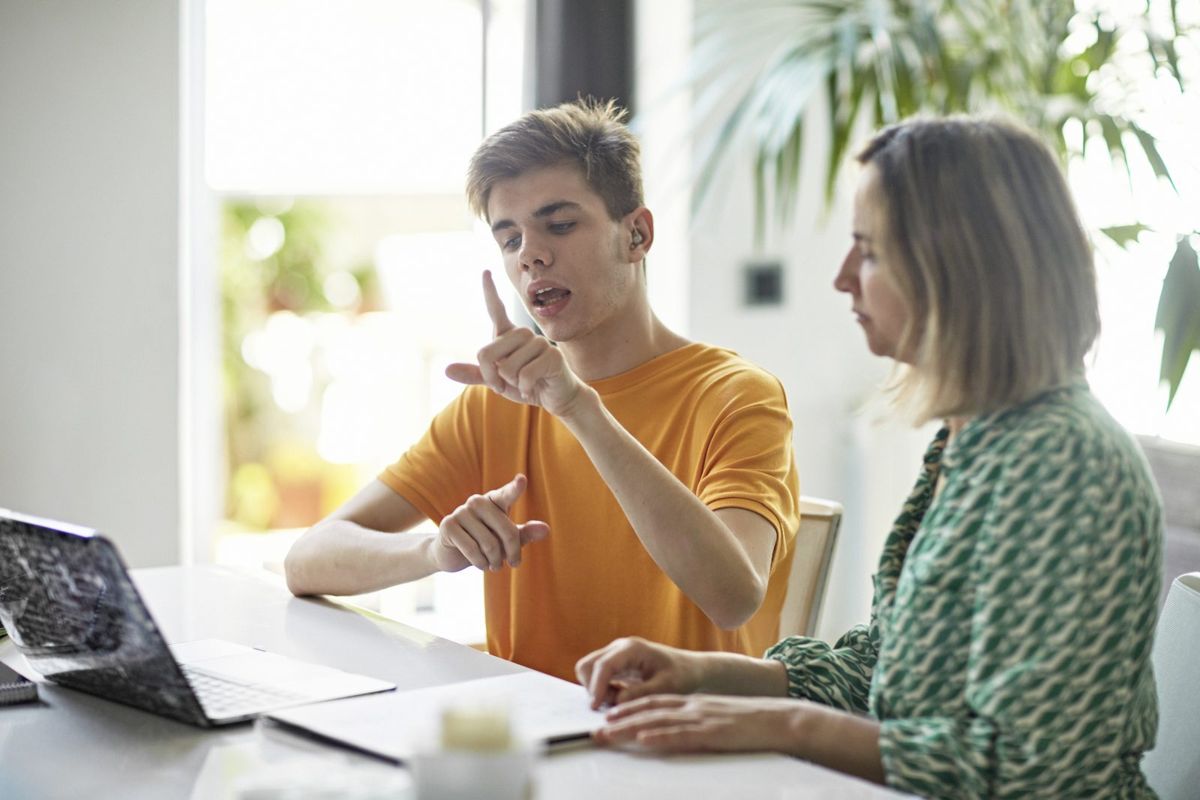 A young man signs in ASL with a coworker at a desk.