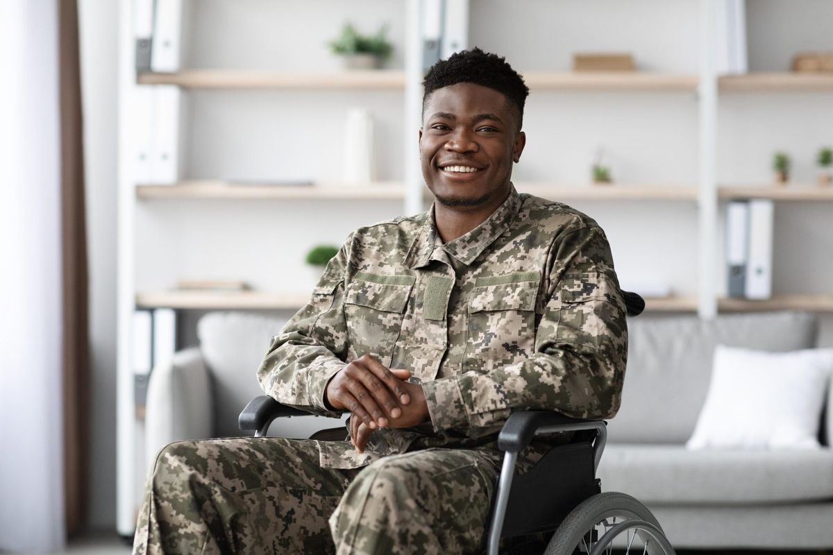 A smiling veteran in uniform sits in a wheelchair at home.