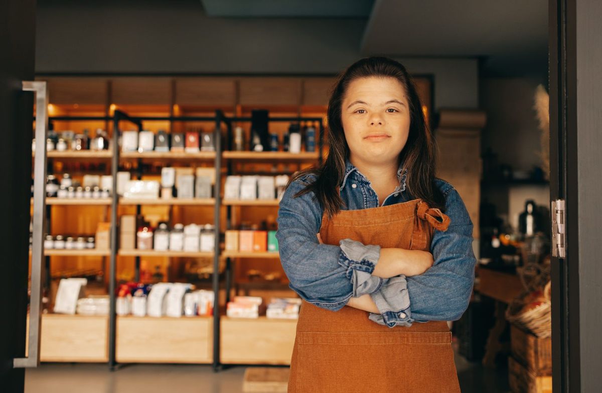 A young woman in an apron stands with arms crossed in her shop.
